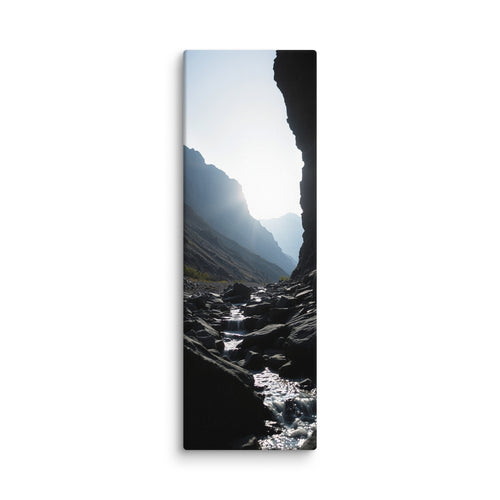 A Thread Through Shadow, tall wrapped canvas print: a bright stream winding through dark shadowed rocks toward hazy mountain ridges.