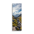 Where The Snow Begins, tall wrapped canvas print: a stream crossing a green alpine meadow toward snow-capped peaks under swirling clouds.