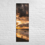Two at the Tide, tall wrapped canvas print: two figures on wet sand under a fiery orange sunset over the sea.