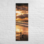 At the Water's Edge, tall wrapped canvas print: two figures on wet sand at the water's edge under a blazing golden sunset.