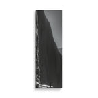 A Place To Stand, tall wrapped canvas print: black-and-white image of a lone figure on a sunlit rocky ledge beside a towering dark cliff.